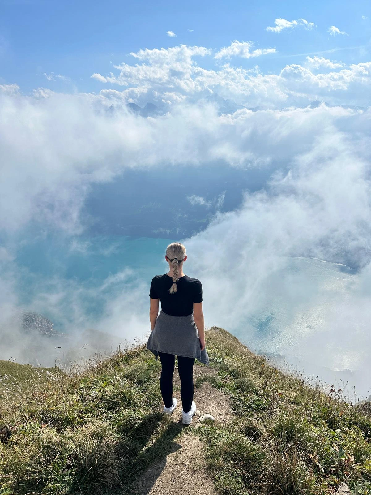 Femme au cheveux blond se tenant au bout d'une colline regardant au loin, ciel bleu avec des nuages blanc